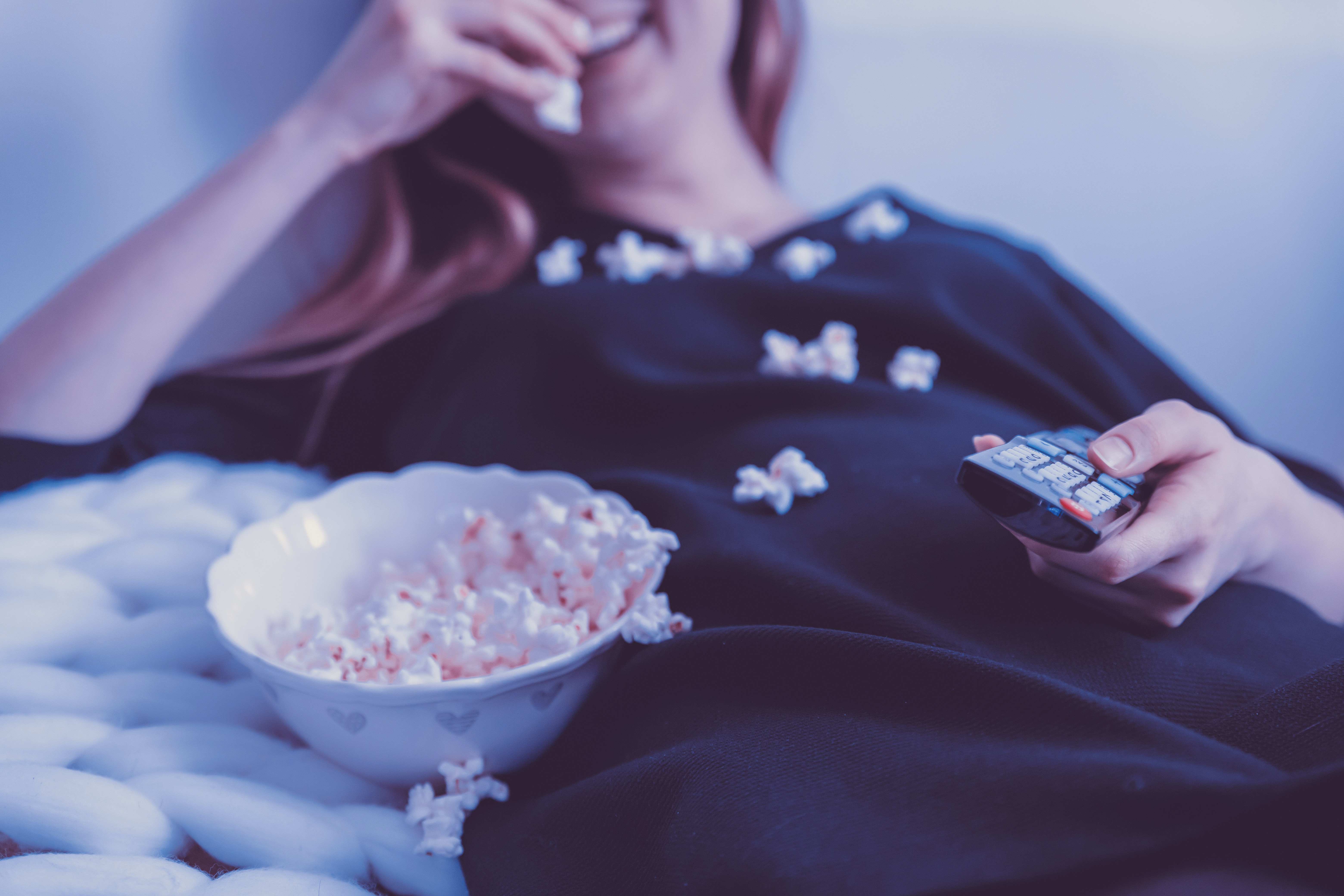 woman lying on bed eating popcorn with a TV remote in hand