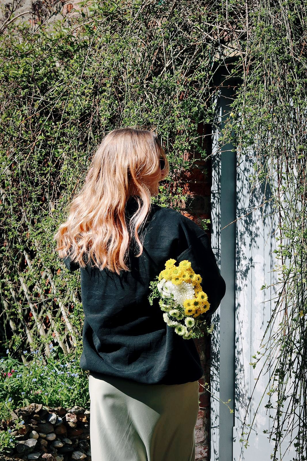 Amy is in an overgrown garden, facing away from the camera holding a bunch of flowers under one arm