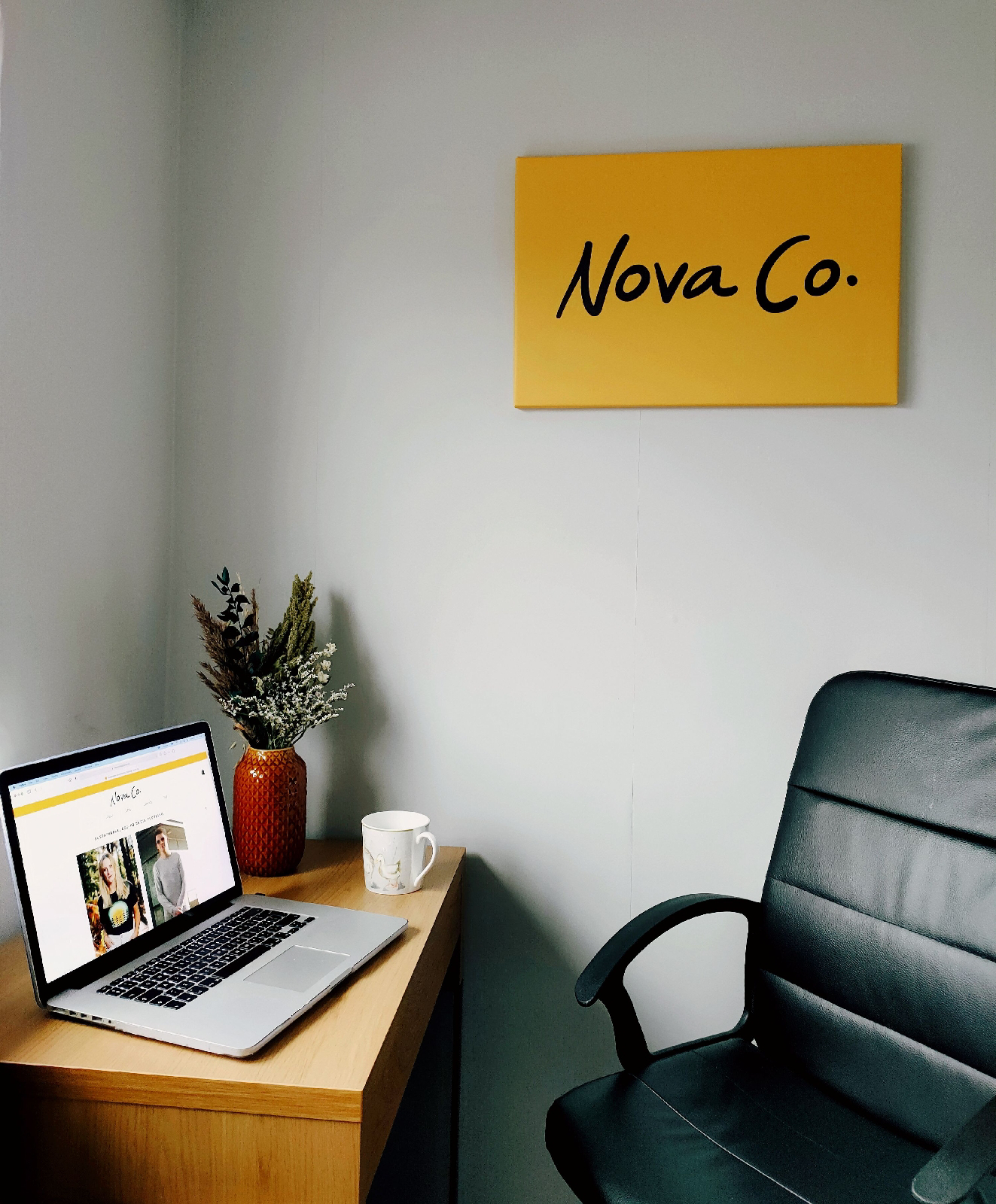 a desk and a chair with a canvas on the wall; there is a laptop and some flowers on the desk