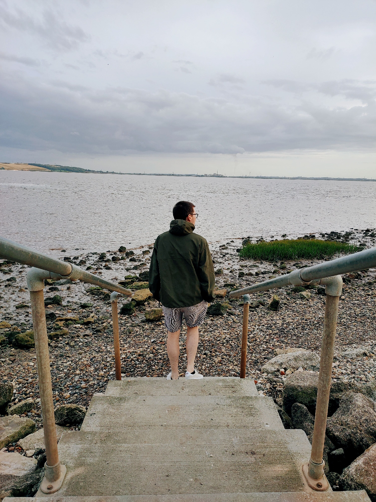 A man stood at the bottom of some steps leading to a pebbled foreshore next to a river.
