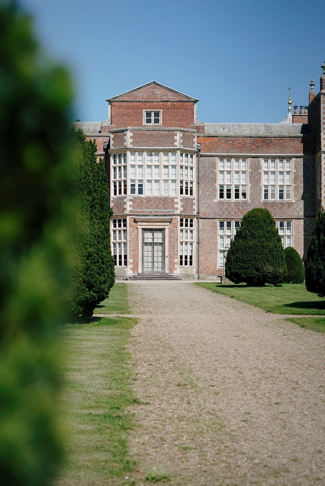 A stately looking country house, view from the path leading up to it with some green hedge in the foreground.