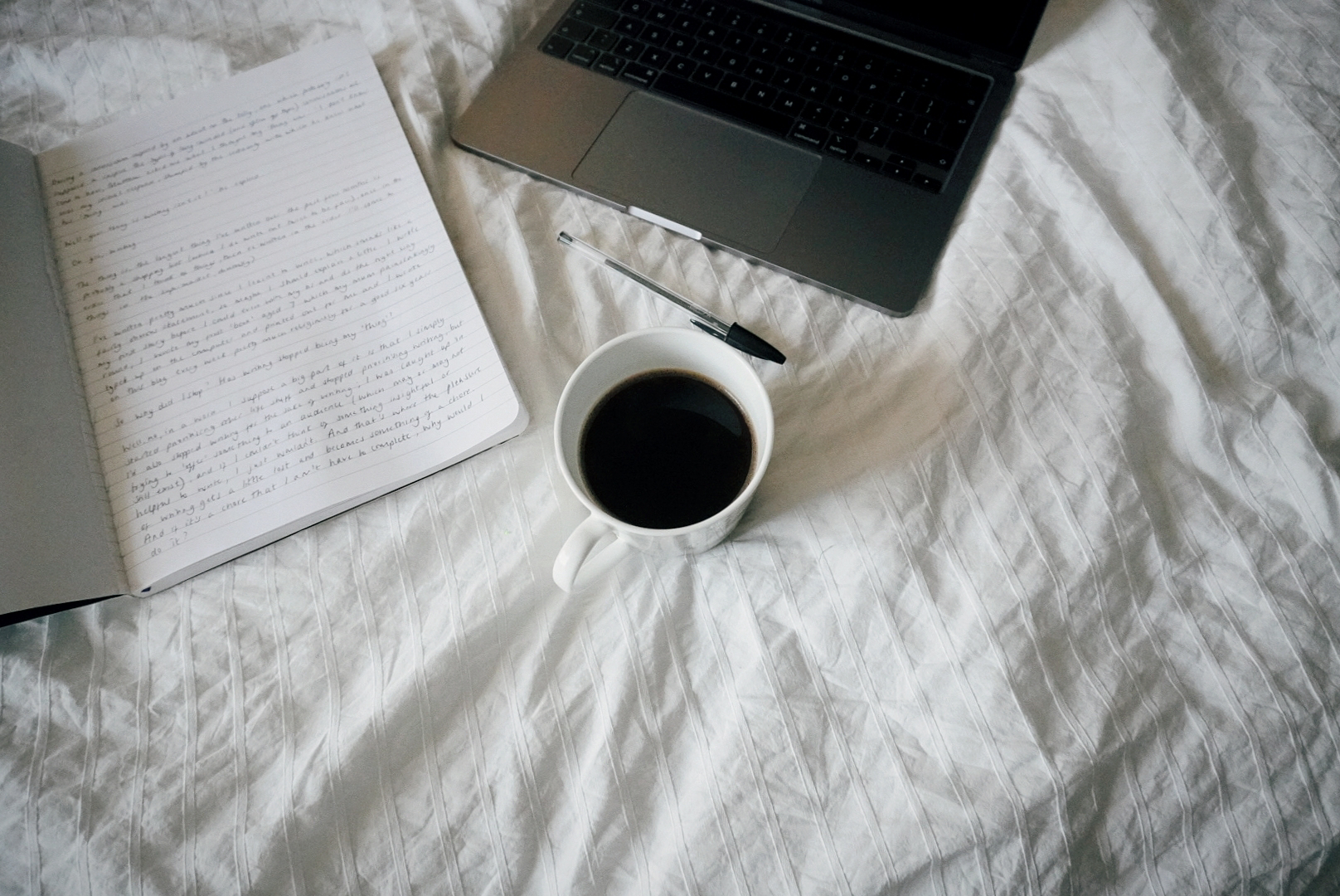 the background is white bedsheets; on the sheets is a laptop, a notebook with some writing in (too small to see) and a mug of black coffee