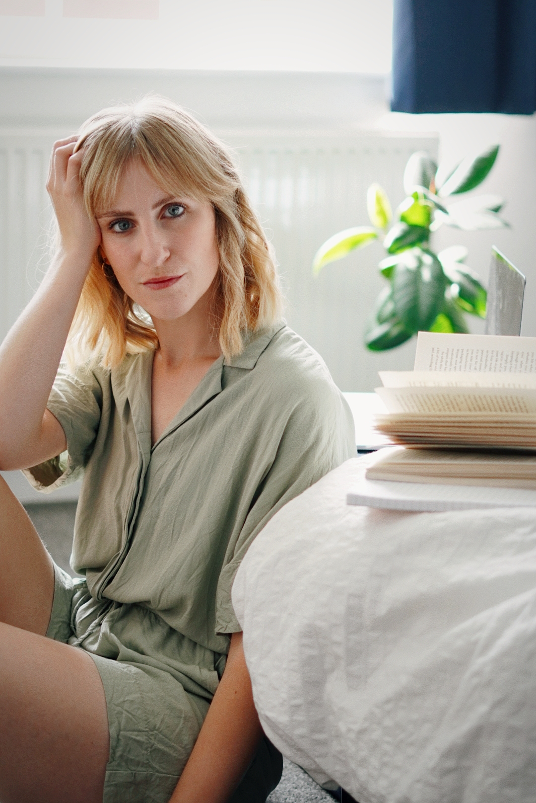 Amy is sitting on the floor next to the bed, wearing a pale green playsuit with her head resting on her hand looking to camera. Next to her are a notebook, book and laptop.