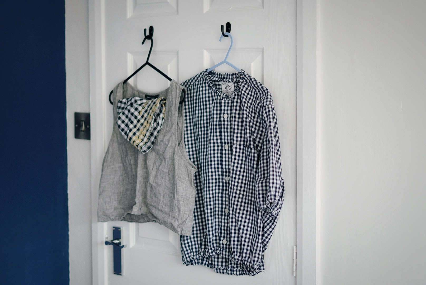 A wider view of the back of a door where a blue gingham blouse, grey cropped top and a black gingham bucket hat are hanging.