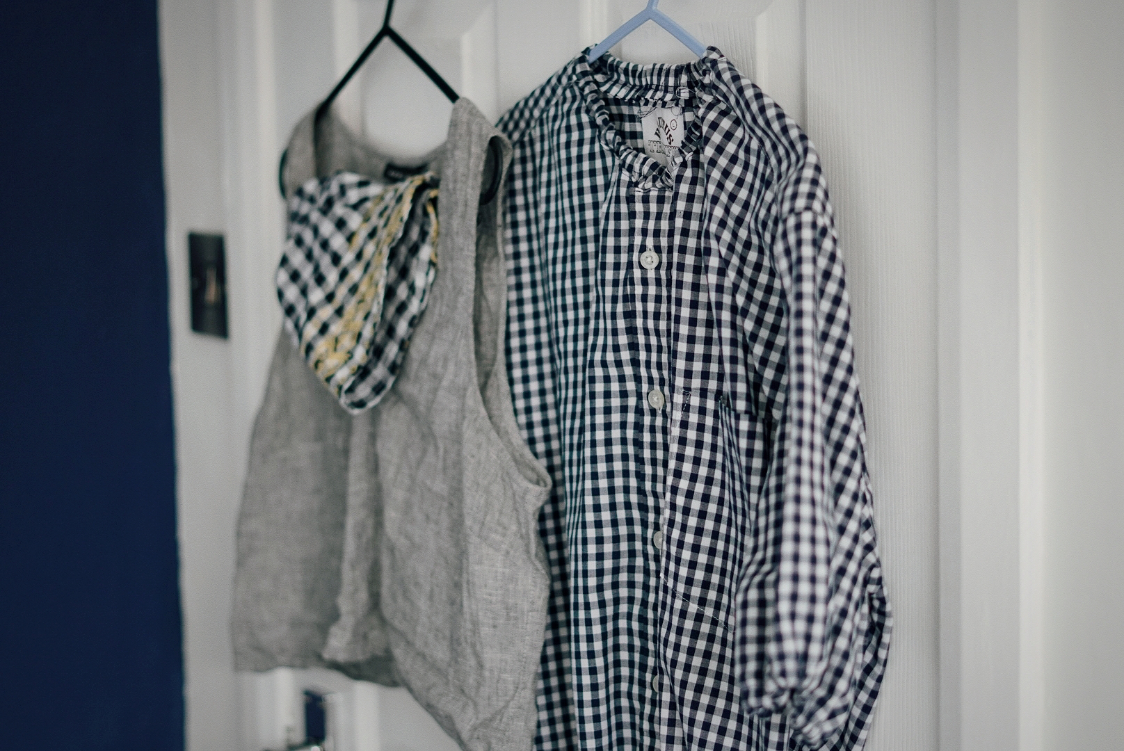 Hung on the back of a door is a navy gingham blouse, a cropped grey top and a black gingham bucket hat.
