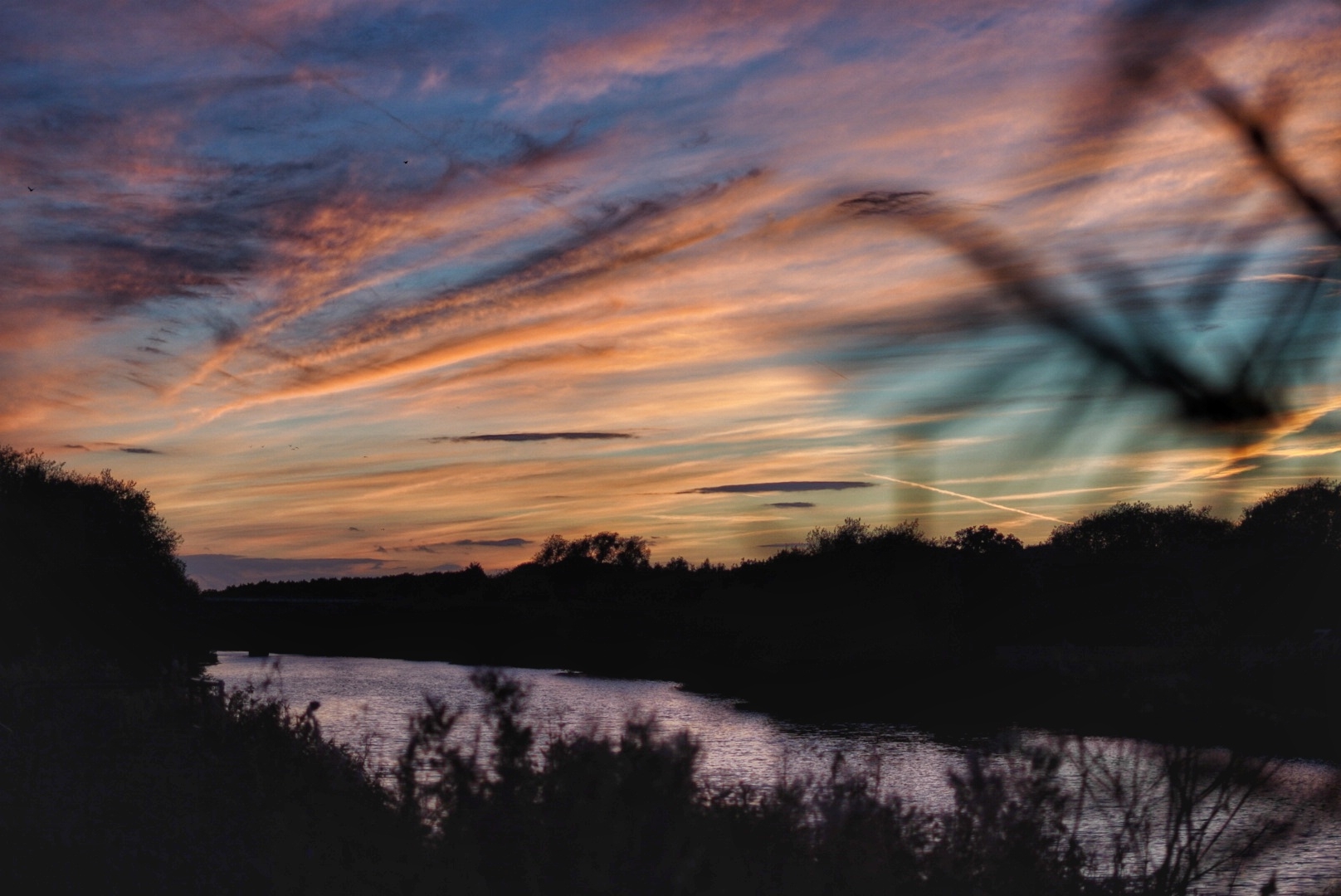 Landscape of a river with banks either side, above is a beautiful sunset; the sky is a mixture of blues, oranges, pinks and purples.