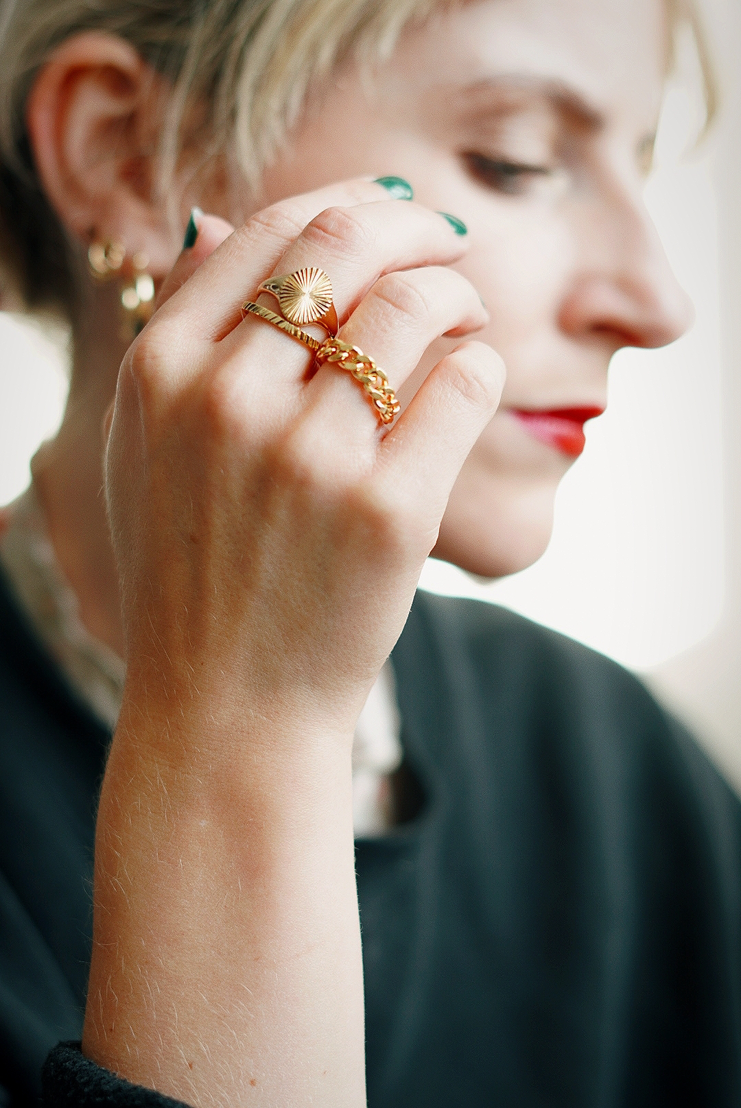 Amy's hand is in focus in front of her face which is not, on her hand are three gold rings; one chain linked, one plain band and one signet ringAmy's hand is in focus in front of her face which is not, on her hand are three gold rings; one chain linked, one plain band and one signet ring. She has green nail varnish on.