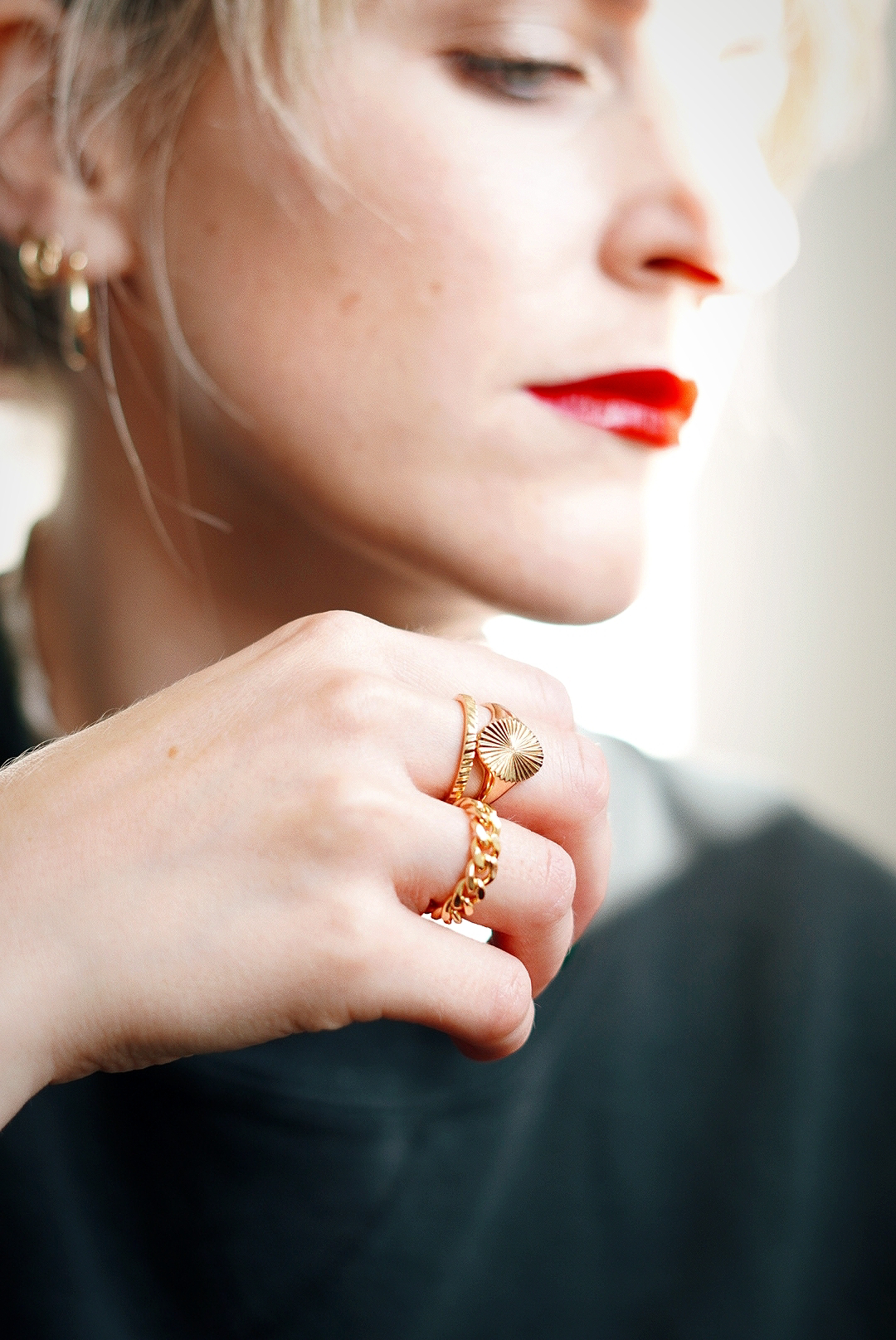 Amy's hand is in focus in front of her face which is not, on her hand are three gold rings; one chain linked, one plain band and one signet ring
