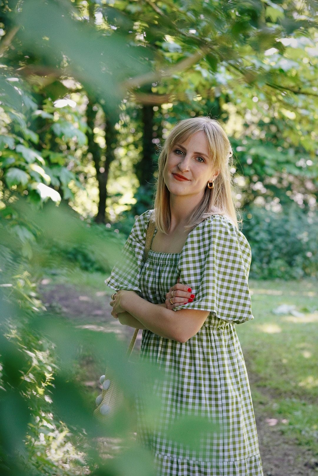 In the foreground are some blurred out leaves framing Amy who is in focus, wearing a green gingham floaty dress.