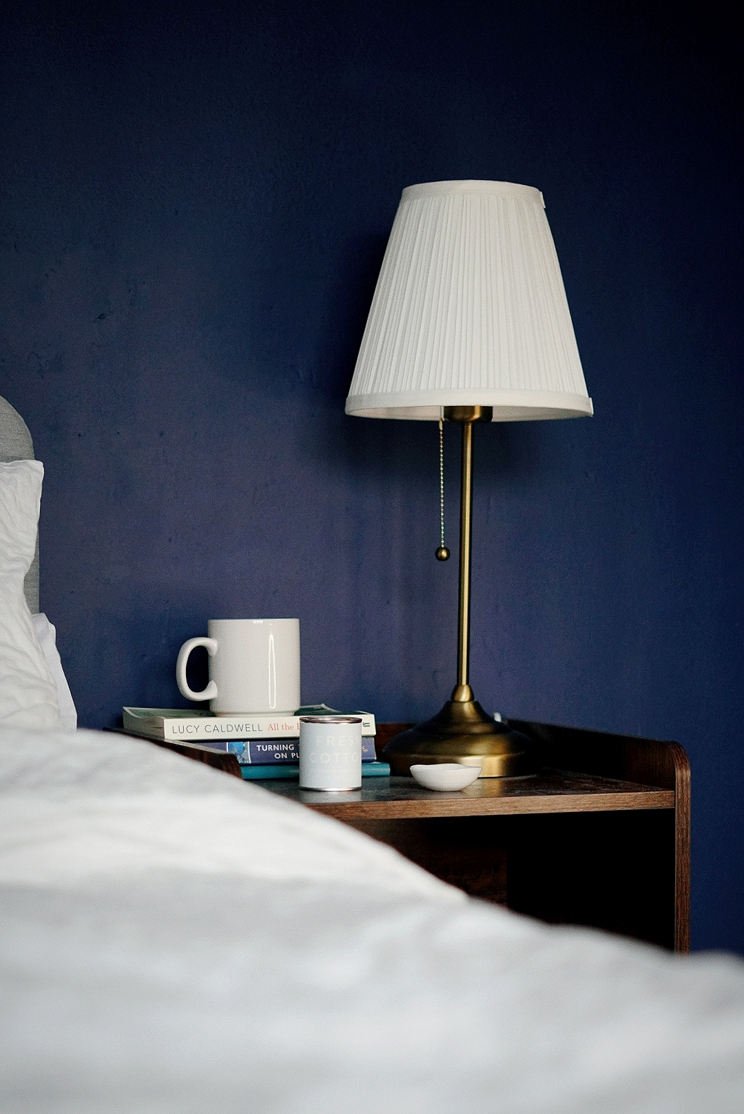 Side view across a white bedspread to a dark wooden bedside table, on top of which is a table lamp, three books with a mug resting on top and a tin candle. The wall behind is dark blue.