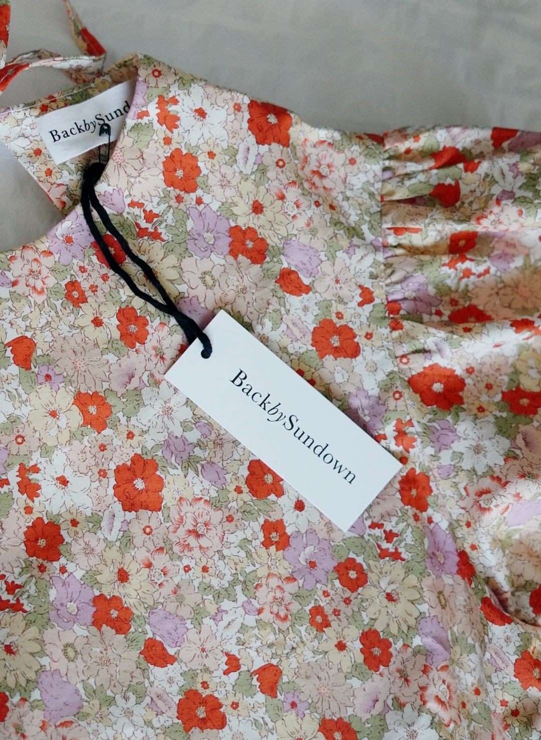 the top half of a floral dress (pinks, greens and oranges) lying on a bed, with the tag saying 'Back By Sundown' poking out of the front