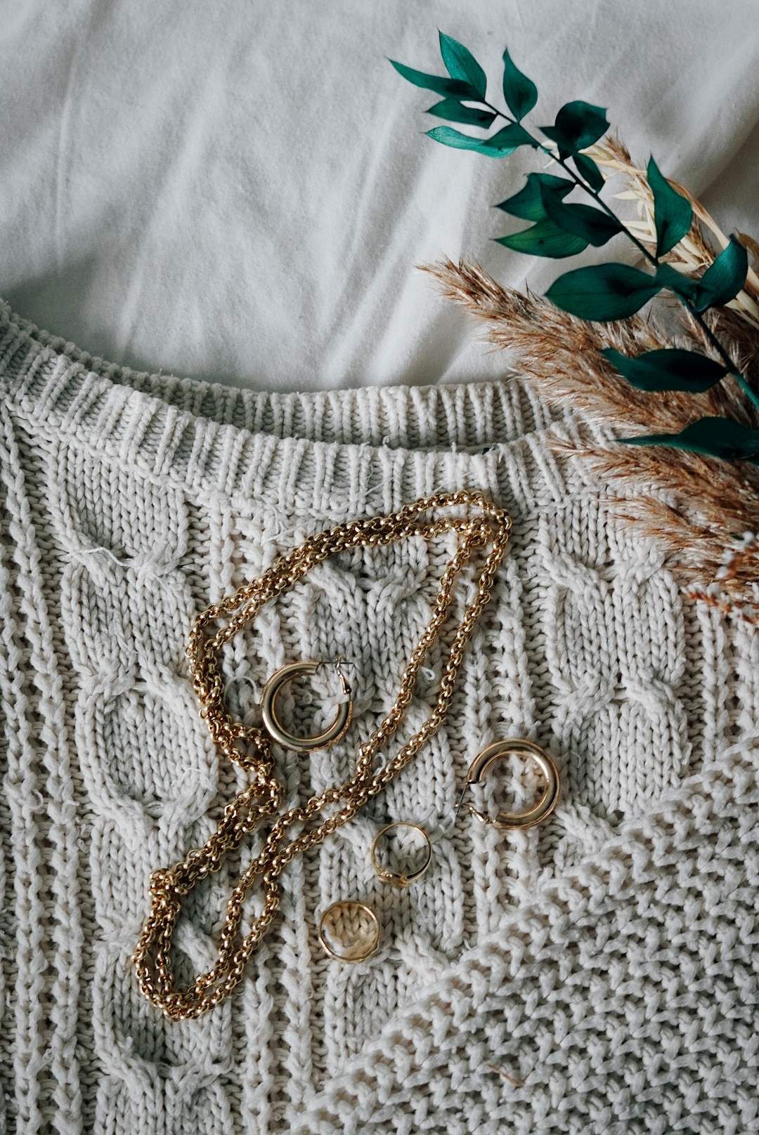 a cream knitted jumper laid on a bed, dried flowers to one side and some simple gold jewellery (a chain and some hoop earrings) lying on top
