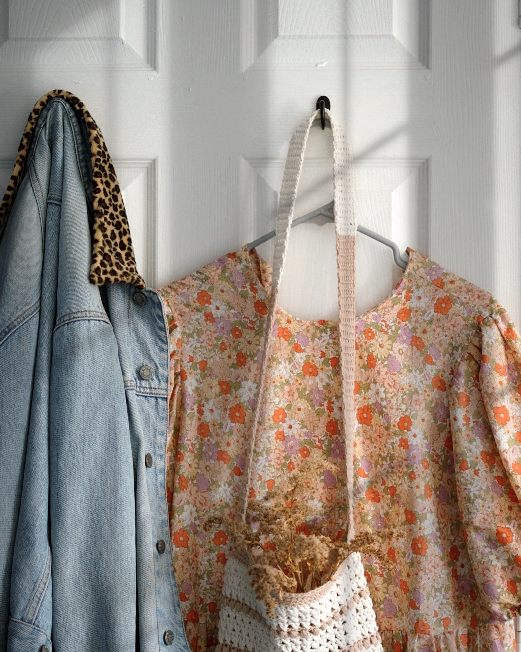 the back of a door with two hooks; on one is a peach floral dress on a hanger and a crochet bag, on the other is a light denim jacket with a leopard print collar