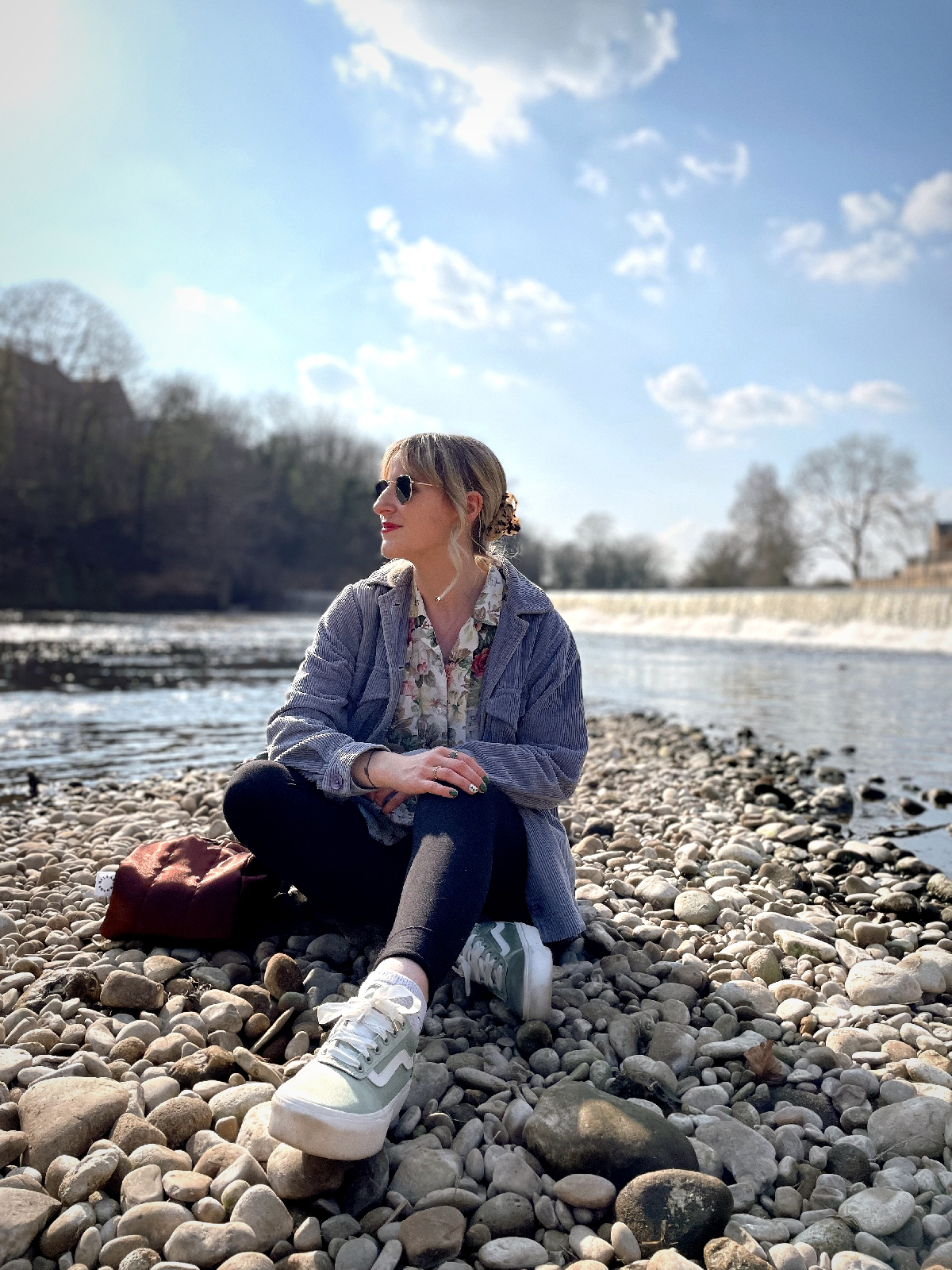 Amy is sitting on some pebbles in front of a stream, the sun hitting the water so it's glistening.