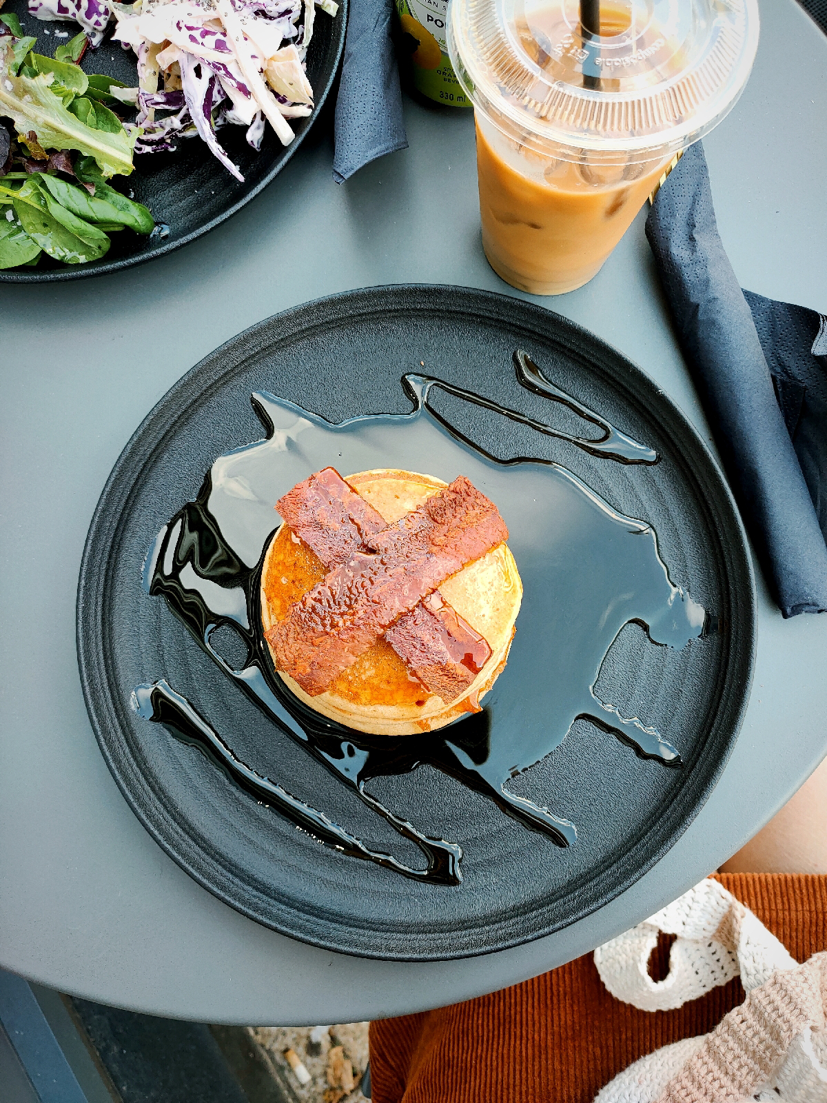 birds eye view of an outdoor table with a plate of pancakes drizzled in maple syrup and vegan bacon and an iced coffee