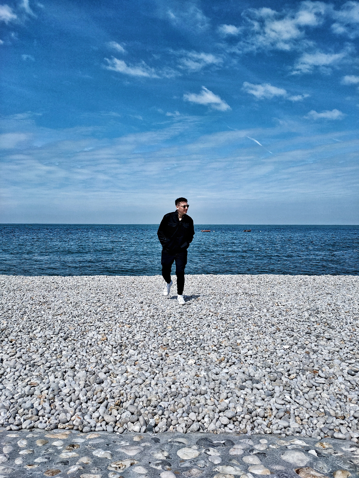 Matthew is walking towards camera across a pebbled beach, looking away. Behind him is the sea and the sky, both very blue