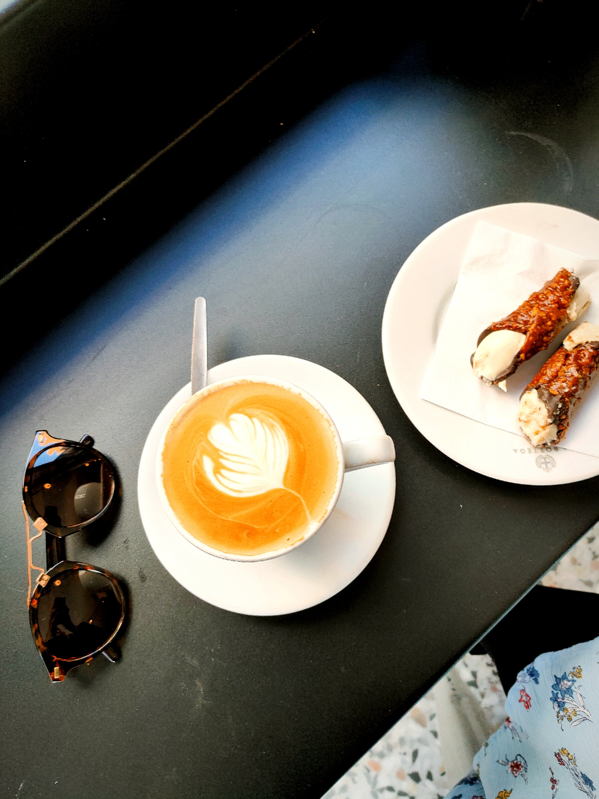 a black surface seen from above, featuring a cup of coffee, a plate with two canolis on it and some sunglasses