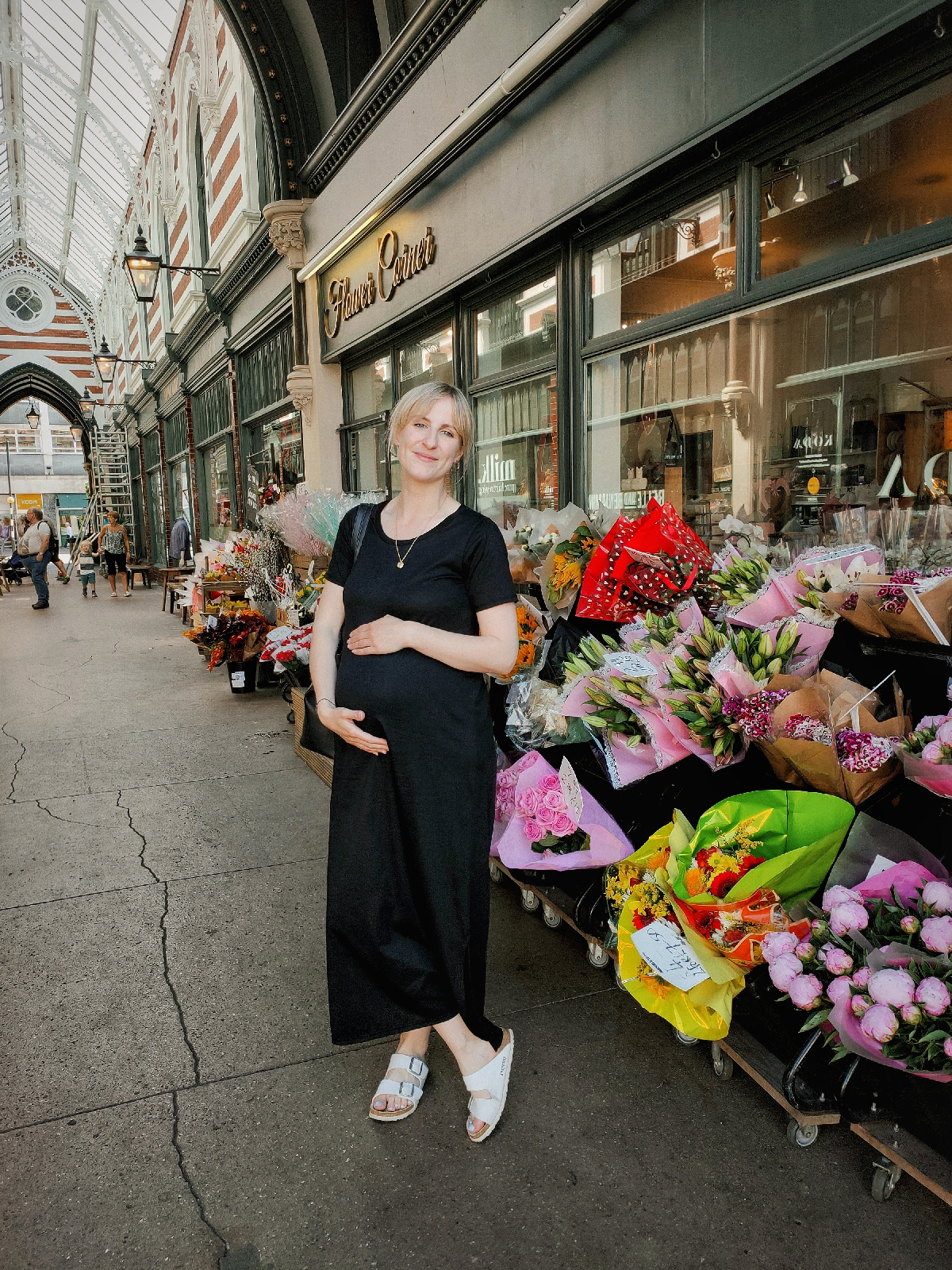 Amy is standing in a shopping arcade in front of a flower shop. She is wearing a black maxi dress and is holding her bump.