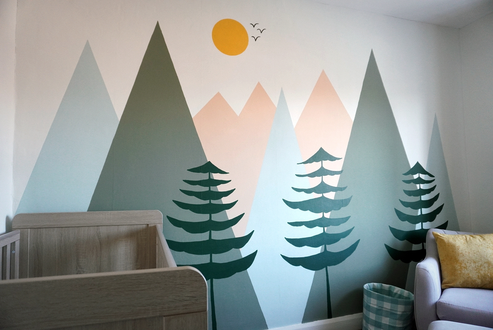 wide angle of a nursery showing a big painted mural on the wall of green, blue and peach mountains, trees and a sun. Also pictured is a wooden cot and an armchair in the corner.