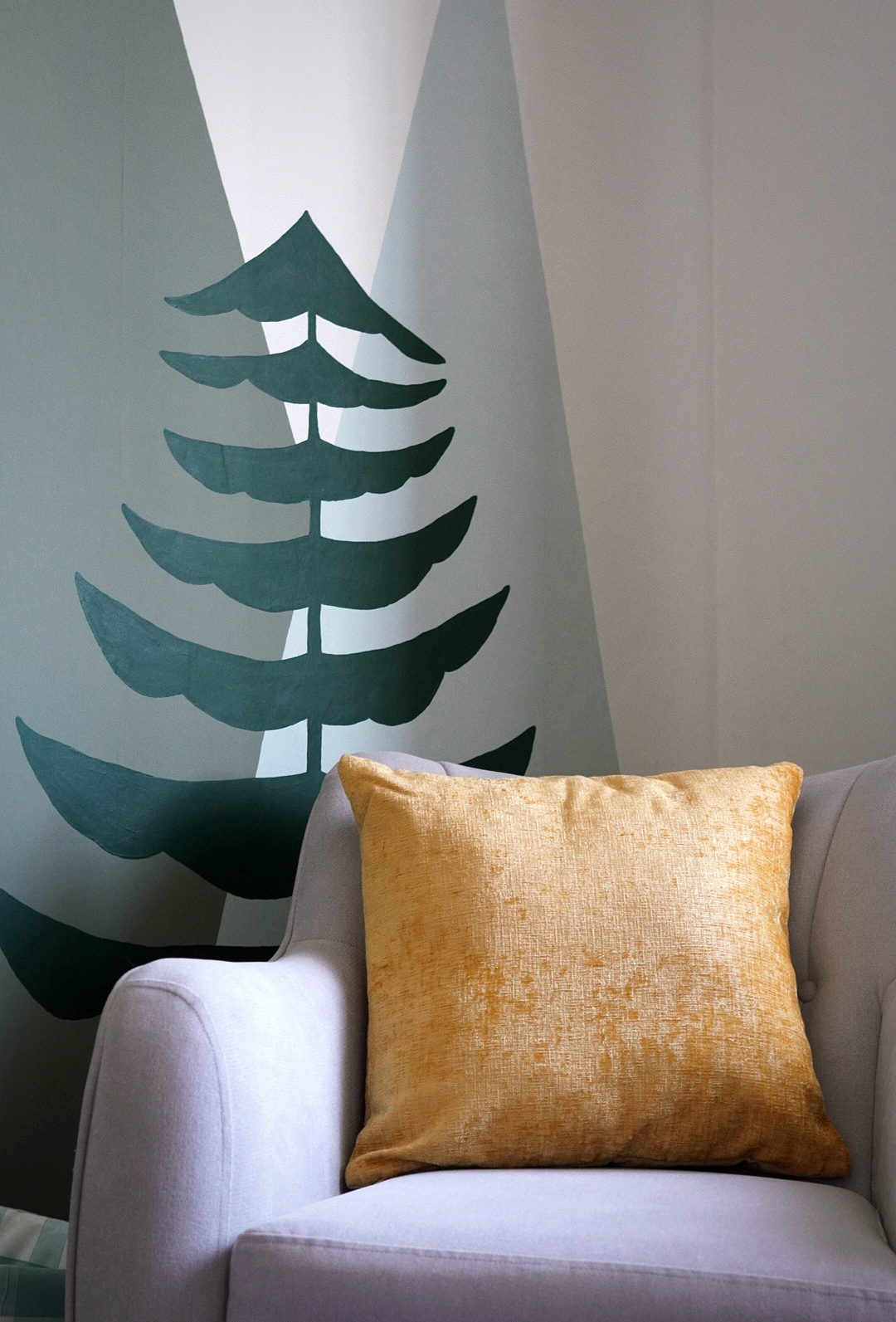 a grey armchair with mustard yellow cushion in the corner of a nursery; behind you can see a painted mural of mountains and trees