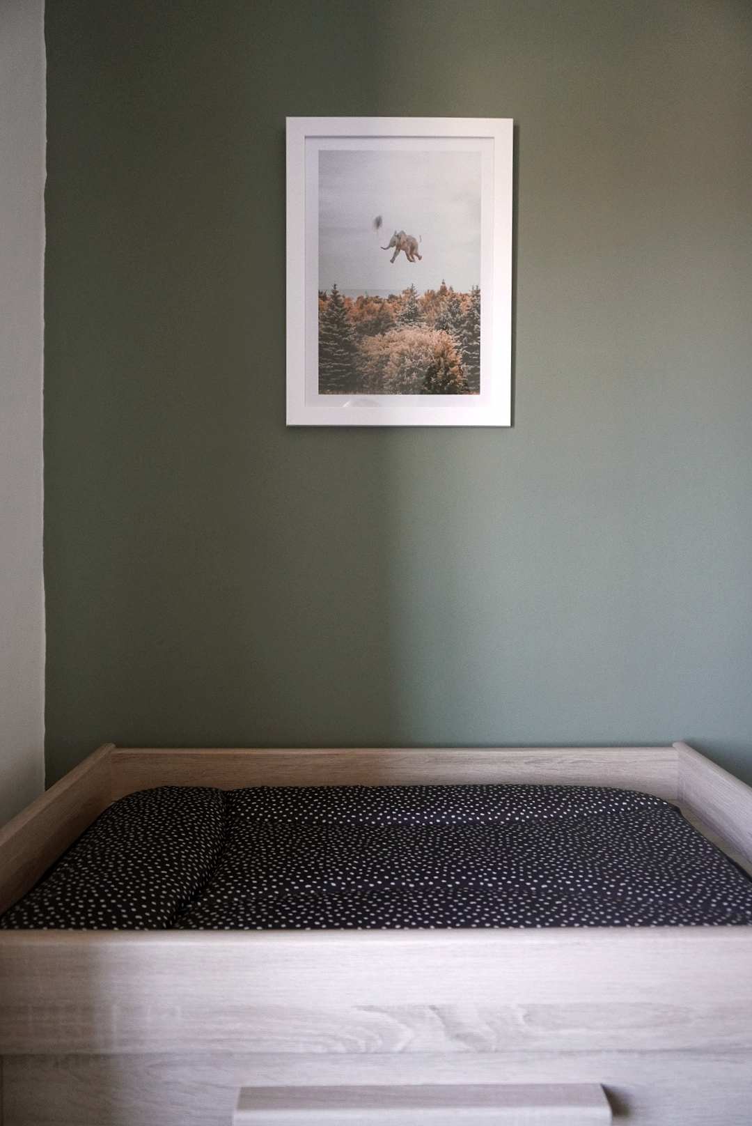 the top of a baby changing table on drawers with a print hanging on the wall behind. The print is of an elephant flying above a forest
