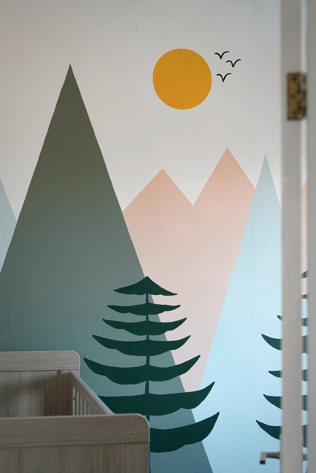looking through a doorway into a nursery; to the foreground is a wooden cot, the wall behind is painted with a mural of green, blue and peach mountains, trees and a sun