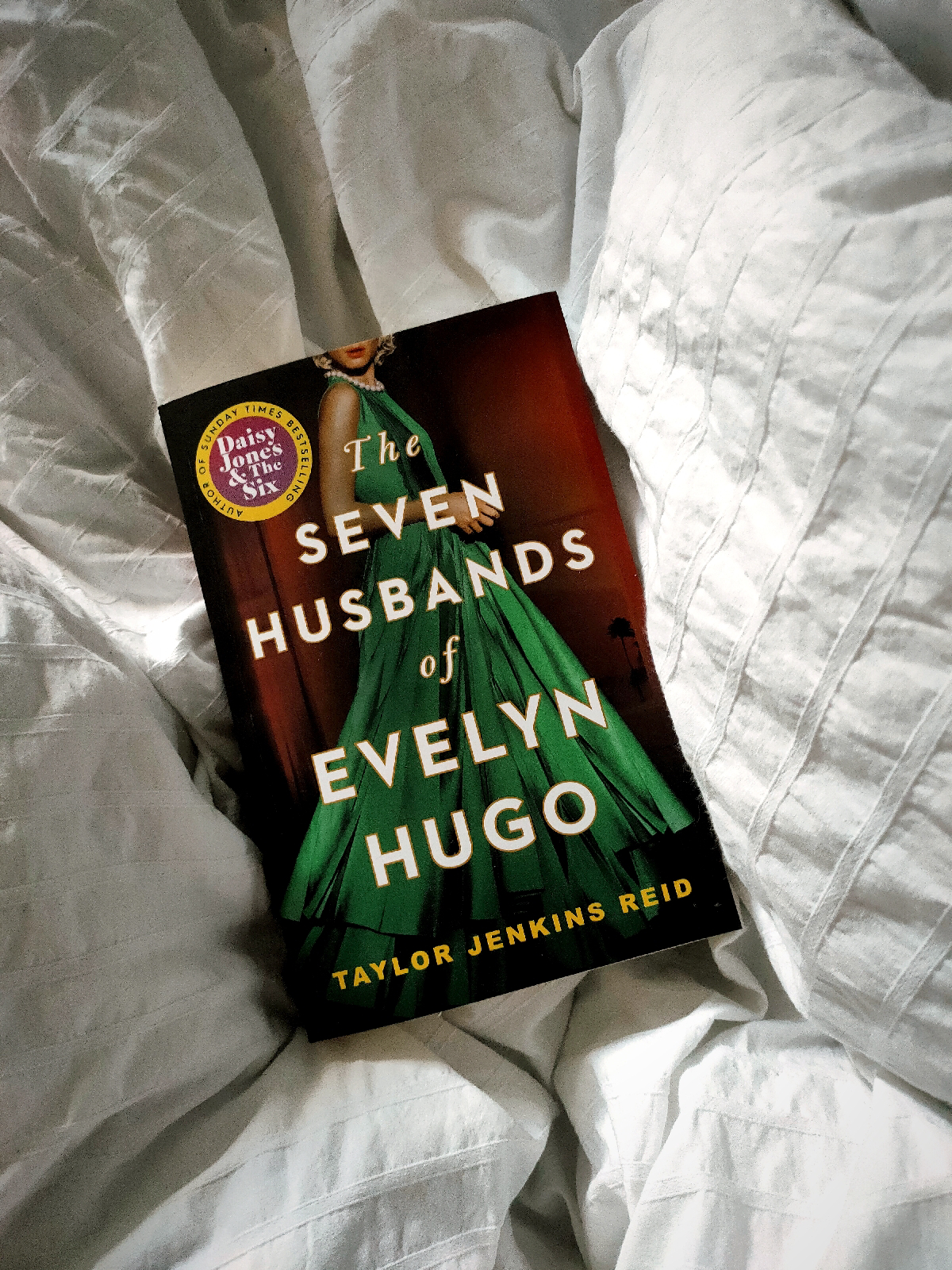 The book 'The Seven Husbands of Evelyn Hugo' featuring an anonymous woman dressed in a green ballgown on the cover is laid on white bedsheets