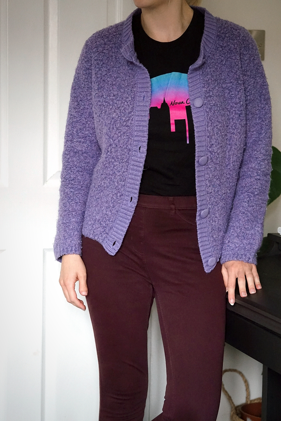 Amy is standing, wearing burgundy skinny jeans, a black t-shirt with a city silhouette design and a lilac cardigan