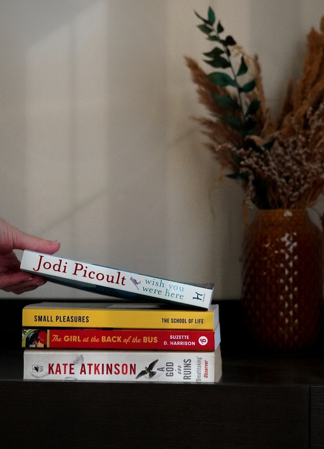 a pile of four books on a dark wooden chest of drawers with dried flowers in the background