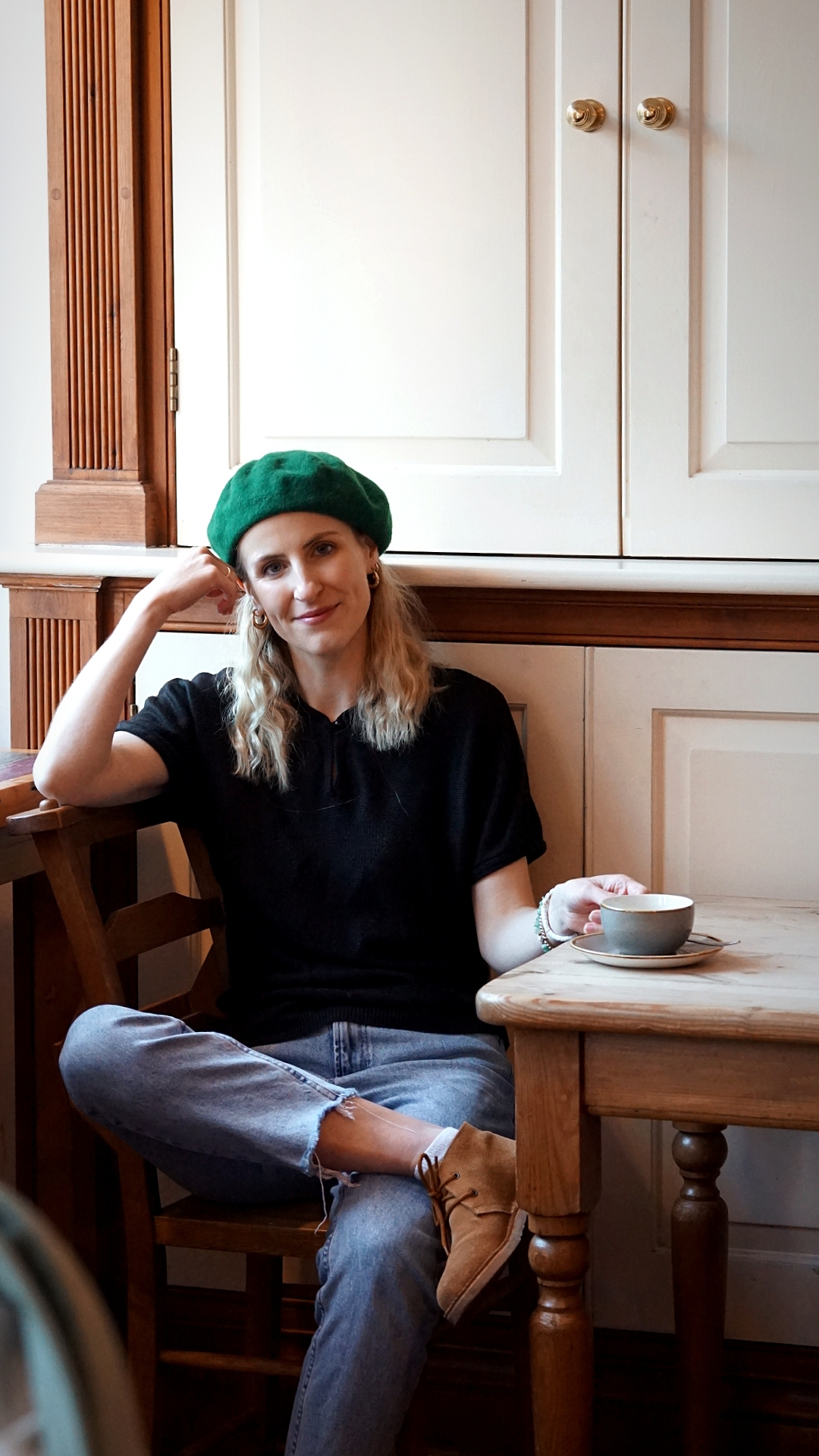 Amy is sitting in a coffee shop looking to camera. She is wearing a black short sleeved jumper, light blue jeans and a green beret.