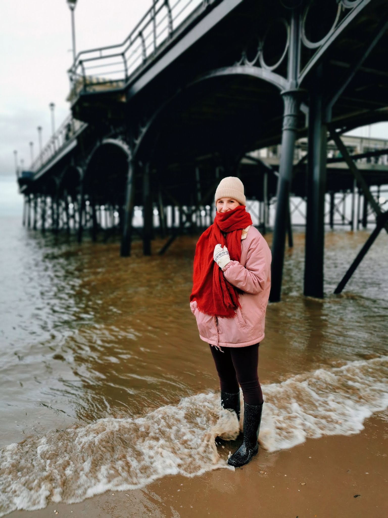 Amy is stood on the beach underneath a huge pier with water lapping at her wellies. She's wearing a pink coat and a red scarf.