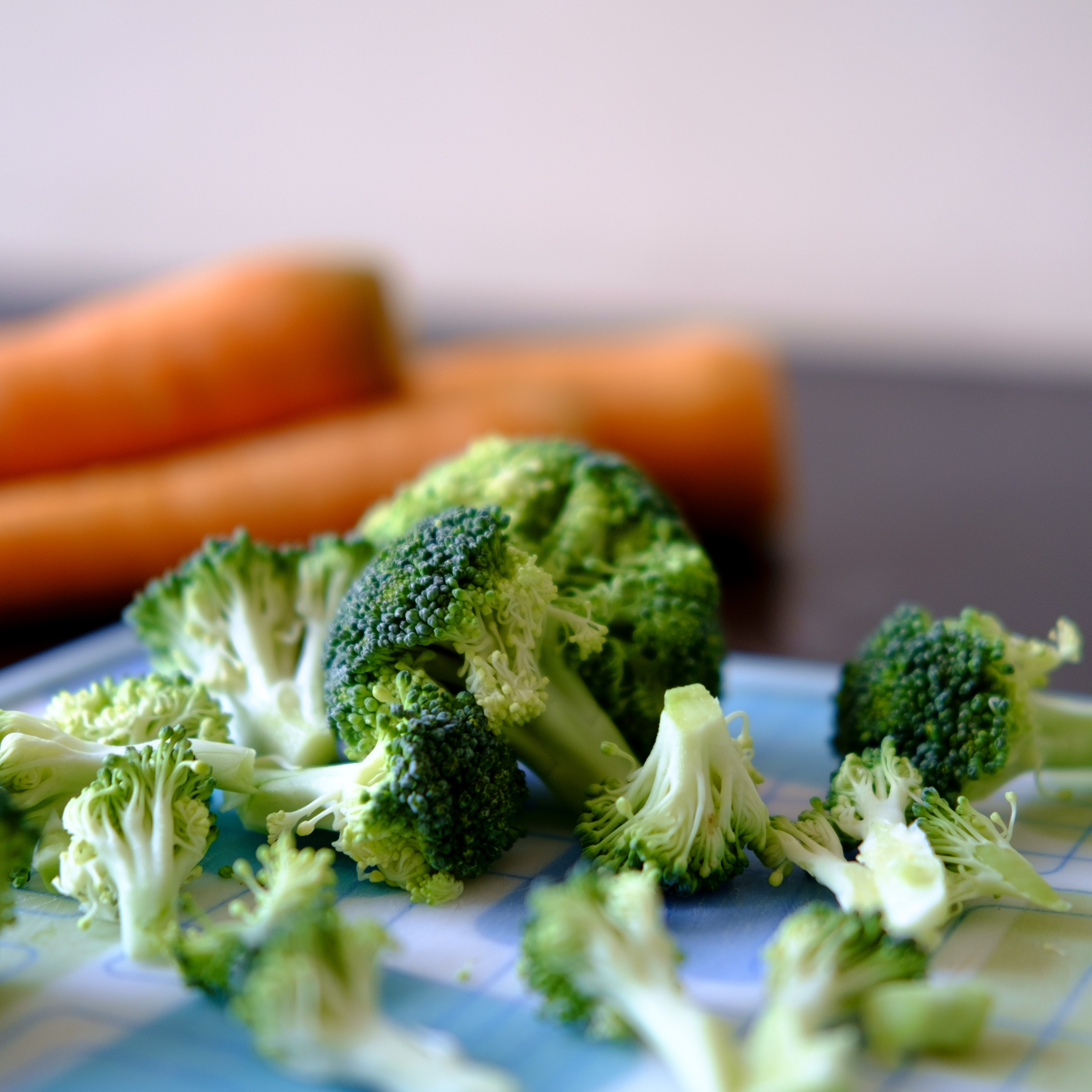 close up of some brocolli on a chopping board, with carrots in the background out of focus