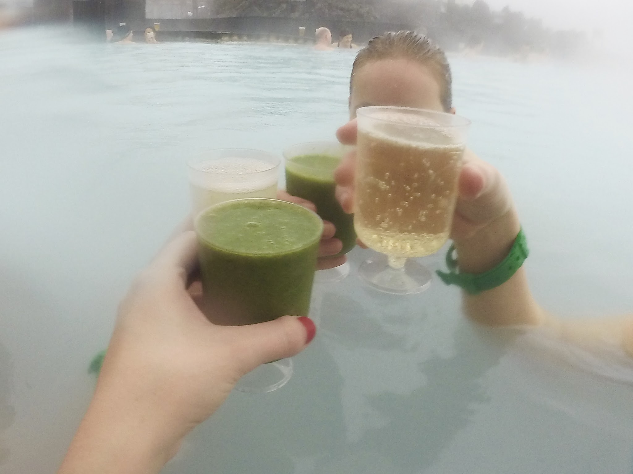 two glasses of prosecco clinking together with two glasses of green juice over the steamy blue water in the lagoon