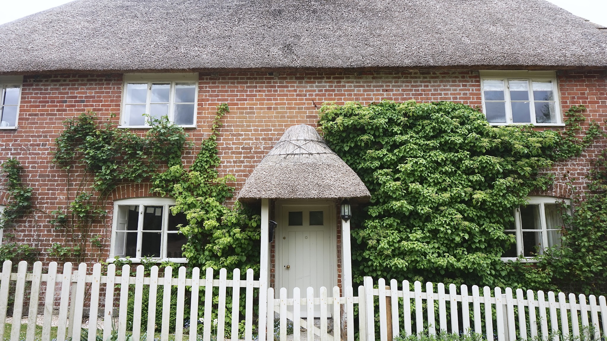 Two-storey brick cottage with a thatch roof, green wooden door and green picket fence