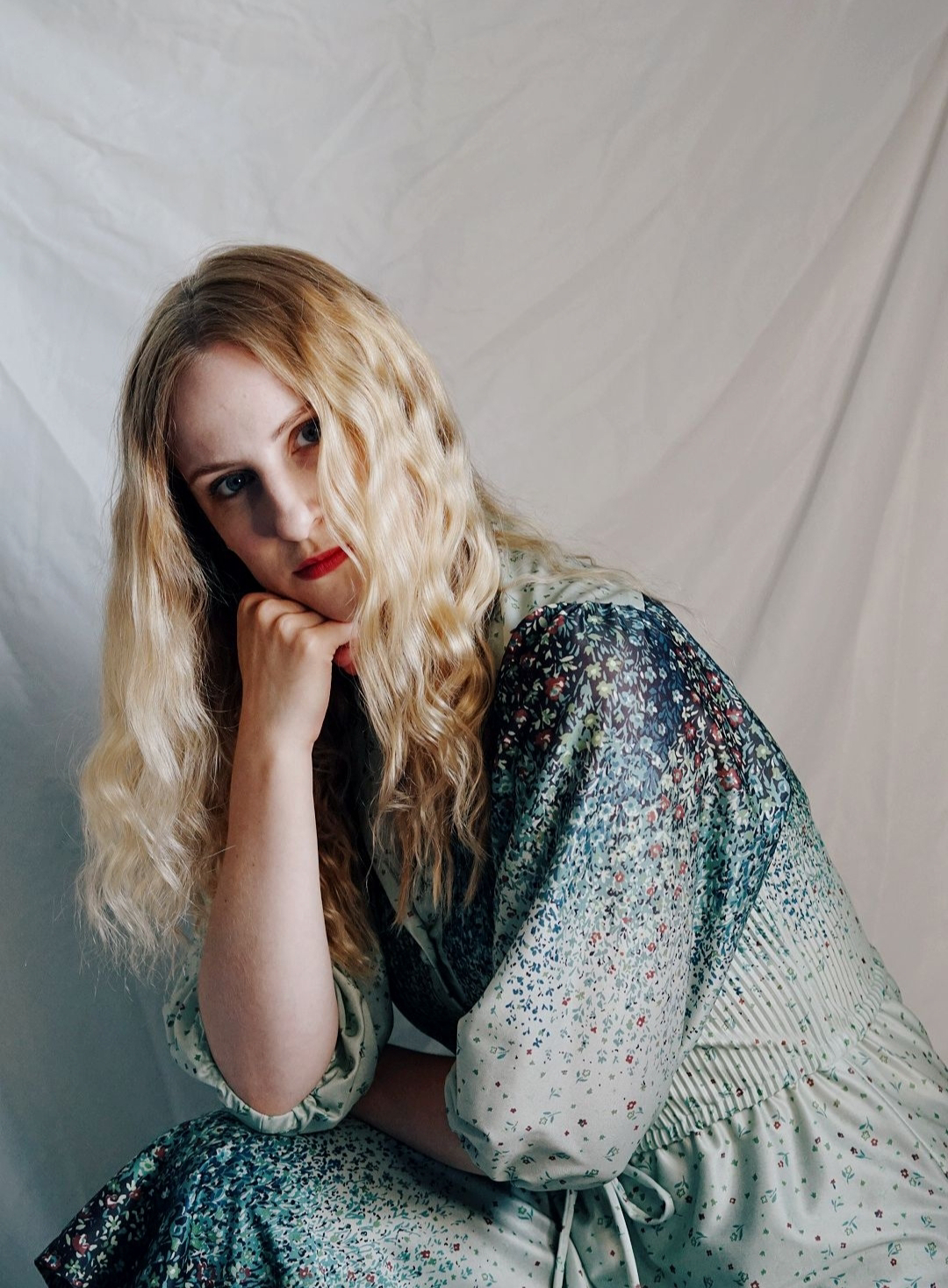 blonde woman knelt down on one knee, looking at the camera sideways on through wavy hair. Wearing a vintage floral silk maxi dress, blue and green.
