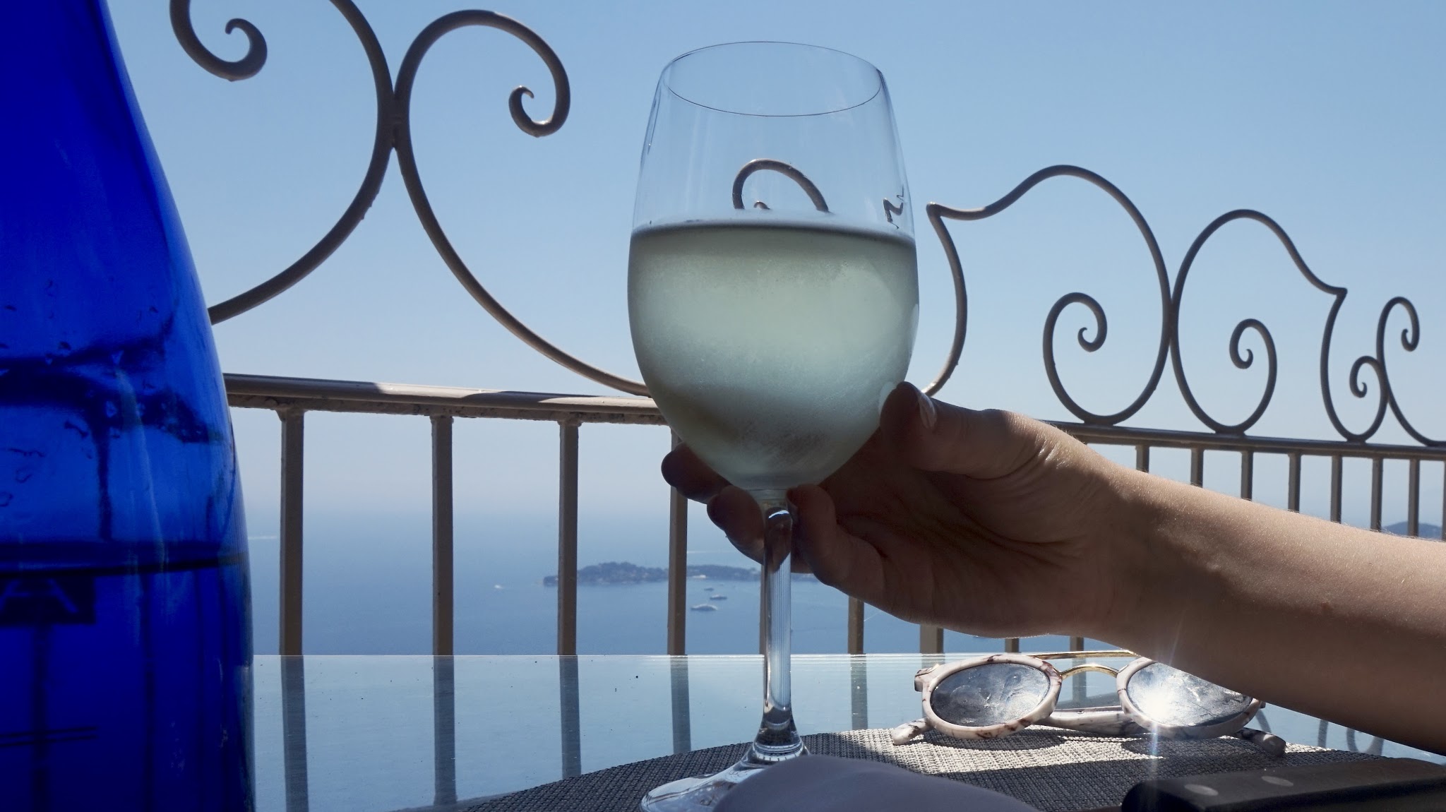 woman holds a glass of white wine in front of some railings overlooking the sea
