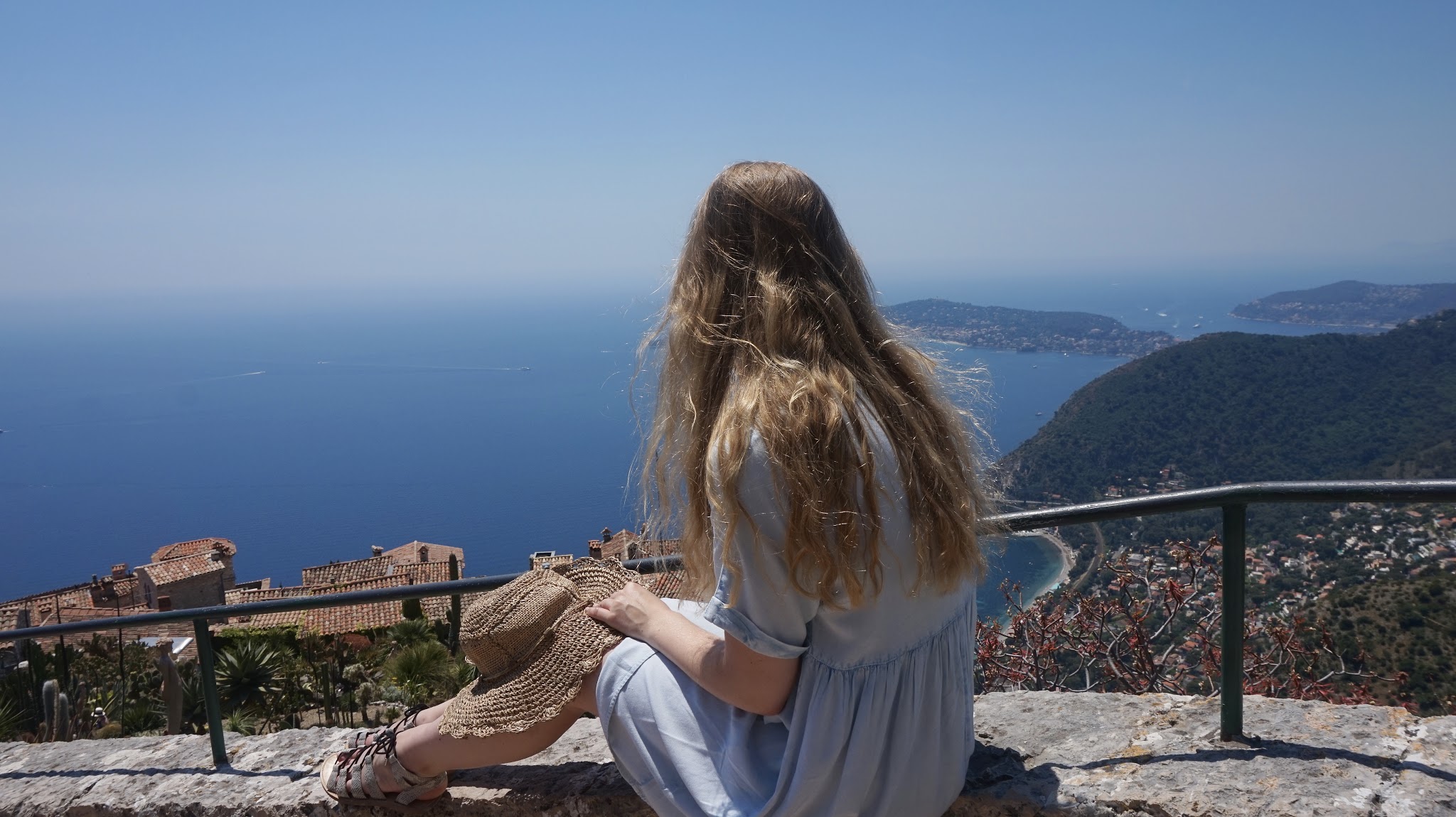 woman sits on a stone ledge overlooking the red rooftops, botanic plants and the sea