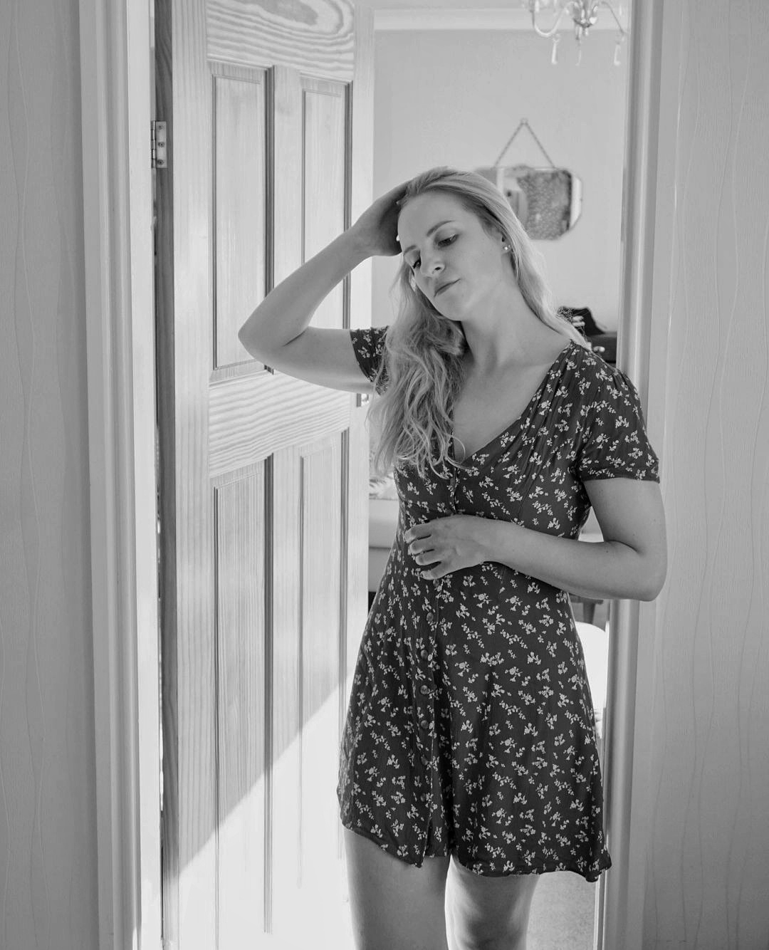 black and white photograph of a woman in a short floral dress standing in a doorway, running her hand through her hair