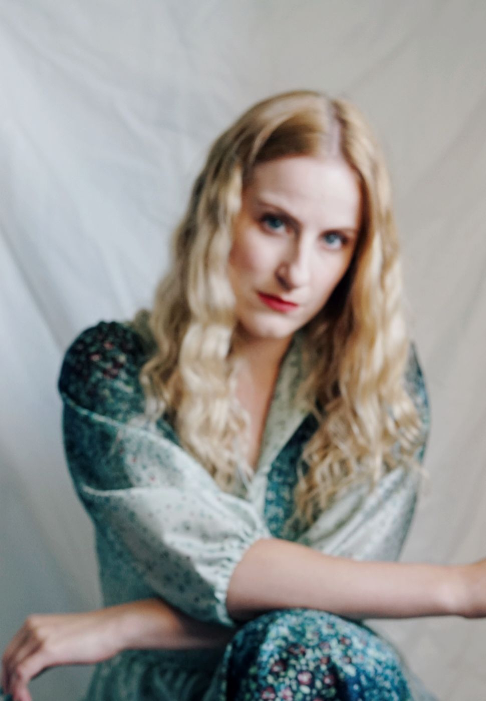 out of focus photograph of woman with wavy blonde hair kneeling facing camera, wearing vintage blue and green floral maxi dress